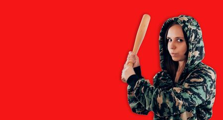 Portrait of young woman in camouflage sweatshirt with bat in hands. Aggressive bully in hood with baseball bat on red background.の写真素材