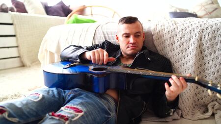 A young man with a tattoo on his chest plays the guitar in the bedroom. Hipster learn to play the...
