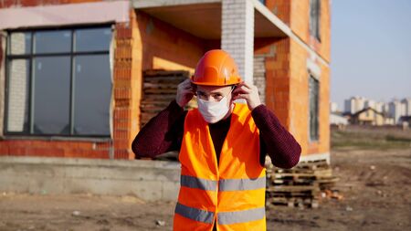 Male construction worker in overalls putting on medical mask on face on construction area.