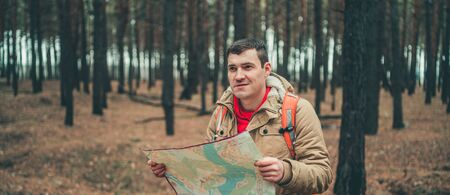 A traveling man with map in woods. A portrait of the handsome man with a backpack, standing near a tree in a cold weather.の写真素材