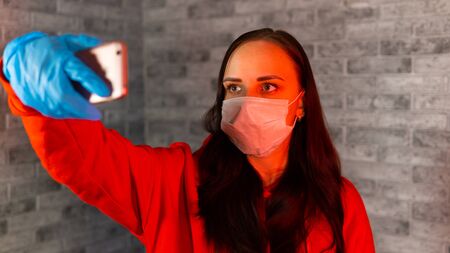 Young woman in medical mask and gloves taking selfie on smartphone, standing on gray brick background. Portrait of female spending time weekend at home and photographing of herself on mobile phone.の写真素材
