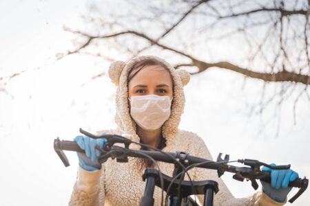 Portrait of young woman in medical mask and gloves with bicycle against sky. Adult female covering face to protect yourself from diseases on walk. Concept of threat of coronavirus epidemic infection.の写真素材
