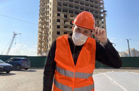 Portrait of male construction worker in medical mask and overalls on background of house under construction.の写真素材