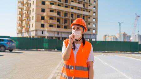 Female construction worker in overalls and medical mask coughing into fist on background of house...