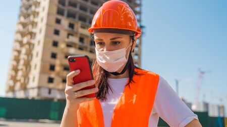 Female construction worker in overalls and medical mask flipping through news about coronavirus in mobile phone on background of house under construction. Concept of threat of infection.の写真素材
