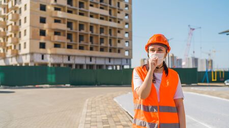 Female construction worker in overalls and medical mask coughing into fist on background of house...