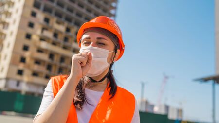 Female construction worker in overalls and medical mask coughing into fist on background of house under construction. Concept of threat of coronavirus epidemic infection.の写真素材