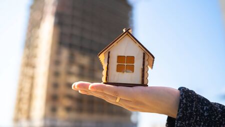 Close up of small wooden house in woman's hand on background of house under construction. Concept of purchasing new apartmentの写真素材