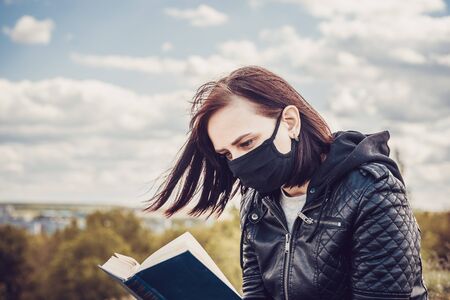 Close up of young woman in black medical mask and casual clothes sitting on hill and reading book on cloudy day. Adult female resting alone in countrysideの写真素材