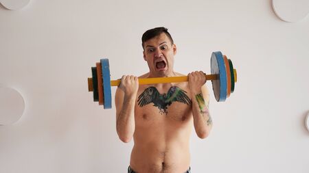 Young man with naked body and tattoo on chest lifting multicolored barbell hard against white...