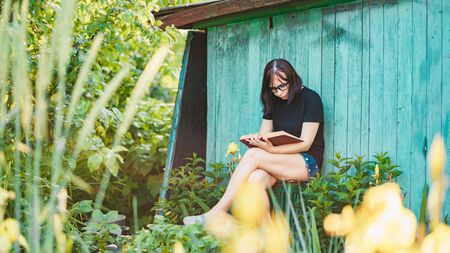 Close up of young woman with glasses reading book in garden. Female resting in nature, enjoying her leisure time.の写真素材