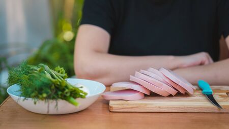 Young woman cutting sausage on wooden board outdoors. Women's hands cutting sausage goods with knife on chopping board.の写真素材