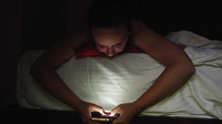 A young woman is lying on the bed and flipping through the various news in a mobile phone before...
