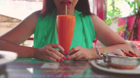 Adult lady sipping fresh fruit beverage through straw while sitting at table in street restaurant on sunny dayの写真素材