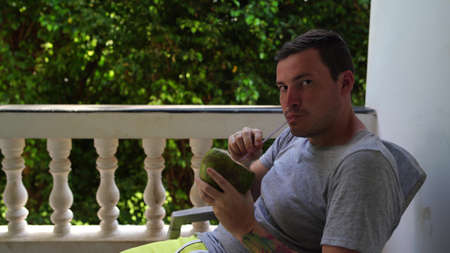 A handsome young man is sitting on a chair and drinking the coconut on the terrace.