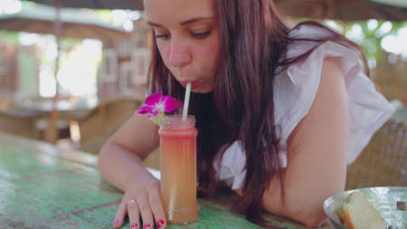 A beautiful young woman drinks a healthy orange smoothie in a tropical cafe.The beautiful girl...