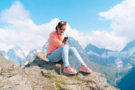 Female traveler using smartphone in mountains. Full body woman sitting on rock and browsing...