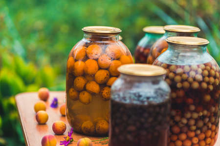 Jars with kompot on table. Sealed glass jars with kompot placed on table near flowers and fruits on summer day in garden.の写真素材