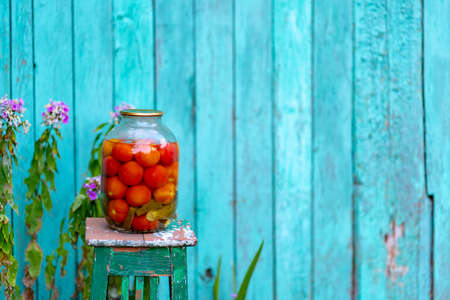 Big glass jar with pickled vegetables on chair on background of old wooden house. Canned tomatoes and cucumbers in sealed jar on street in gardenの写真素材