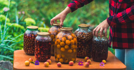 Close up of canned juice with fruits and berries on table in garden. Unrecognizable woman takes...