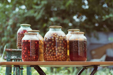 Jars with kompot on table. Sealed glass jars with kompot placed on table near flowers and fruits on summer day in garden.の写真素材