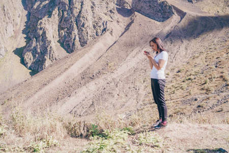 Young woman browsing mobile phone, standing in hilly area. Female traveler stands with smartphone on background of mountain in summertimeの写真素材