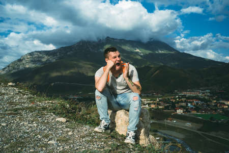Tired traveling man resting on hill. Exhausted male tourist sitting on stone and sweetly yawning...
