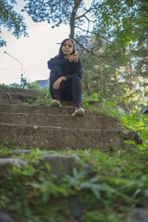 Low angle of young female traveler in activewear sitting on old stone stairs and drinking coffee while resting during walk in green botanical parkの写真素材