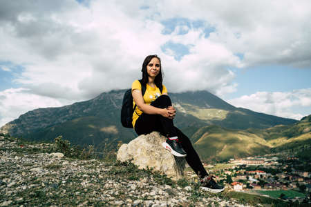Young woman sitting on hill. Dreamy female sitting on altitude and looking at Peaks of magnificent rocks located against bright cloudy sky on sunny day in nature.の写真素材