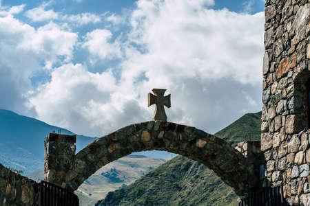 Close up of stone gate with wooden cross on background of mighty mountains and cloudy sky. Entrance...