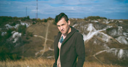Tattooed man with bare torso in cape and jeans stands in hilly terrain. Adult male poses on cloudy day in countryside.の写真素材