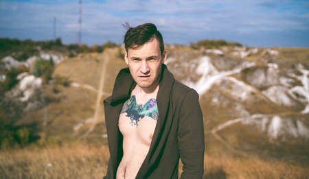 Tattooed man with bare torso in cape and jeans stands in hilly terrain. Adult male poses on cloudy day in countryside.の写真素材