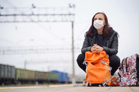 Young woman in medical mask squats on platform, waiting for train. Female passenger in protective mask with backpacks sitting on railroad platform in waiting for train ride during coronavirus pandemic.の写真素材