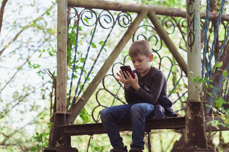 Focused boy using smartphone in park. Serious concentrated school aged boy in casual wear sitting on bench and playing game on mobile phone while spending autumn day in park.の写真素材