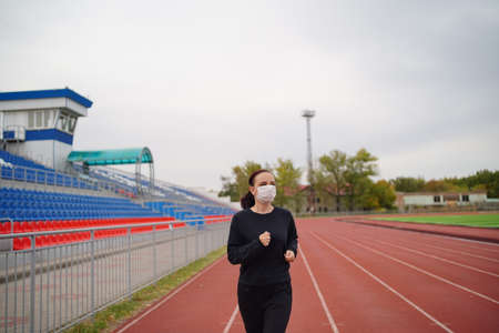 Active woman in protective mask running at stadium. Young fit female in sportswear and protective mask for coronavirus prevention running on red track during outdoor workout at stadium.の写真素材