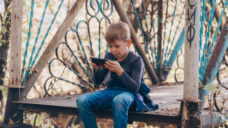 Focused boy using smartphone in park. Serious concentrated school aged boy in casual wear sitting on bench and playing game on mobile phone while spending autumn day in park.の写真素材