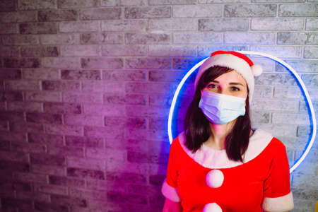 Young woman in medical mask and Santa Claus suit against illuminated wall. Close up of brunette in Christmas hat and protective mask. Concept of safe Christmas celebration during coronavirus pandemic.の写真素材