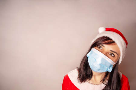 Young woman in medical mask and Santa Claus suit on gray background. Close up of female in Christmas hat and protective mask.Concept of safe Christmas celebration during coronavirus pandemicの写真素材