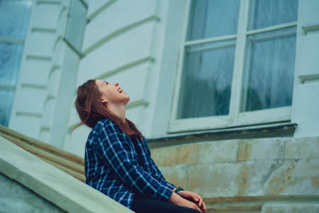 Young woman laughs, sitting on steps of mansion. Side view of pretty smiling female sitting near window.の写真素材