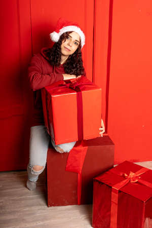 Young attractive woman in Santa Claus hat sits on gifts near red wall. Smiley female sitting on big box of gift. Concept of holidays, gifts and good moodの写真素材