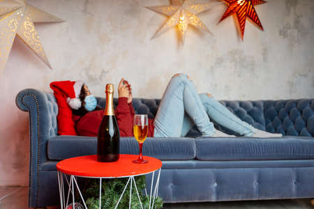 Close up of bottle and glass of champagne on table. Woman in medical mask and Santa Claus hat...