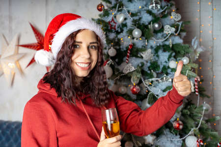 Young woman in Santa Claus hat with glass of champagne standing near Christmas tree. Charming brunette posing at coniferous tree with decorative adornments. Concept of Christmas celebration at homeの写真素材
