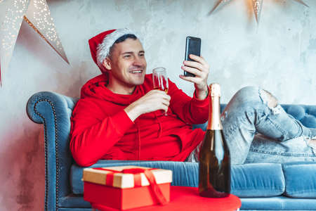 Young handsome man in Santa Claus hat with glass of champagne conducting live broadcast with friends on smartphone. Happy guy celebrating Christmas online on internet and lying on couch at homeの写真素材
