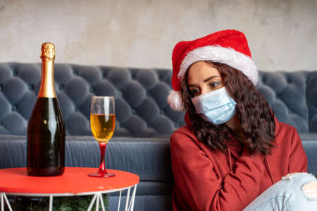 Young woman in medical mask and Santa Claus hat sitting on floor near sofa in room. Alone charming brunette in protective mask celebrating of safe Christmas during coronavirus pandemicの写真素材