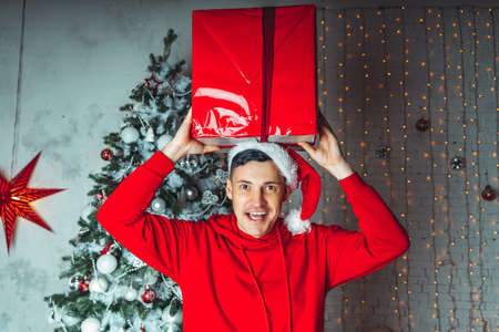 Young handsome man in Santa Claus hat dancing with big gift on background of Christmas tree. Happy guy dances, rejoicing present. Concept of holidays, presents and good moodの写真素材