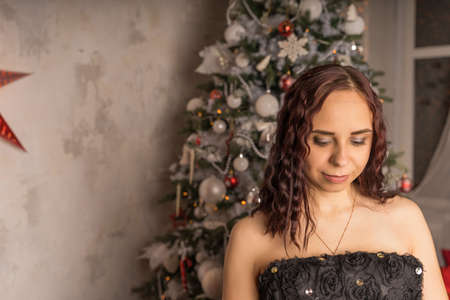 Portrait of charming brunette in black dress near Christmas tree. Young attractive woman posing at coniferous tree with decorative adornments. Concept of Christmas celebration at homeの写真素材