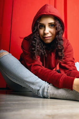 Young woman in hood sitting on floor. Curly brunette poses near red wall.の写真素材