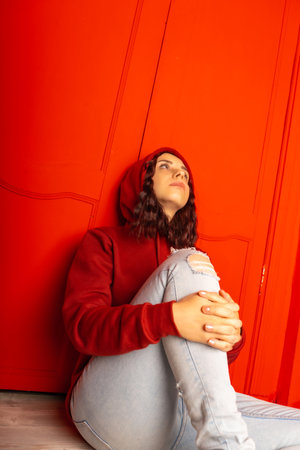 Young woman in hood sitting on floor. Curly brunette poses near red wallの写真素材