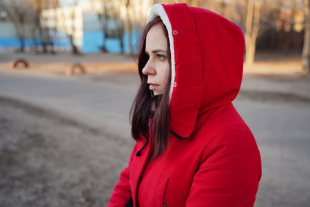 Portrait of young woman in red hood and jacket on street. Adult serious female looks away thoughtfully, standing on fresh air.の写真素材