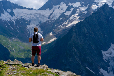 Rear view of man with backpack standing on peak of mountain. Male tourist enjoys beautiful view of mighty mountains in sunny weatherの写真素材
